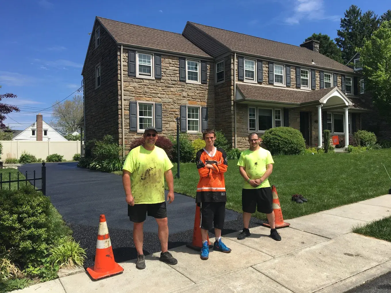 Three crew members in work shirts standing at the end of a freshly sealed driveway in front of a stone colonial, cones on the sidewalk.
