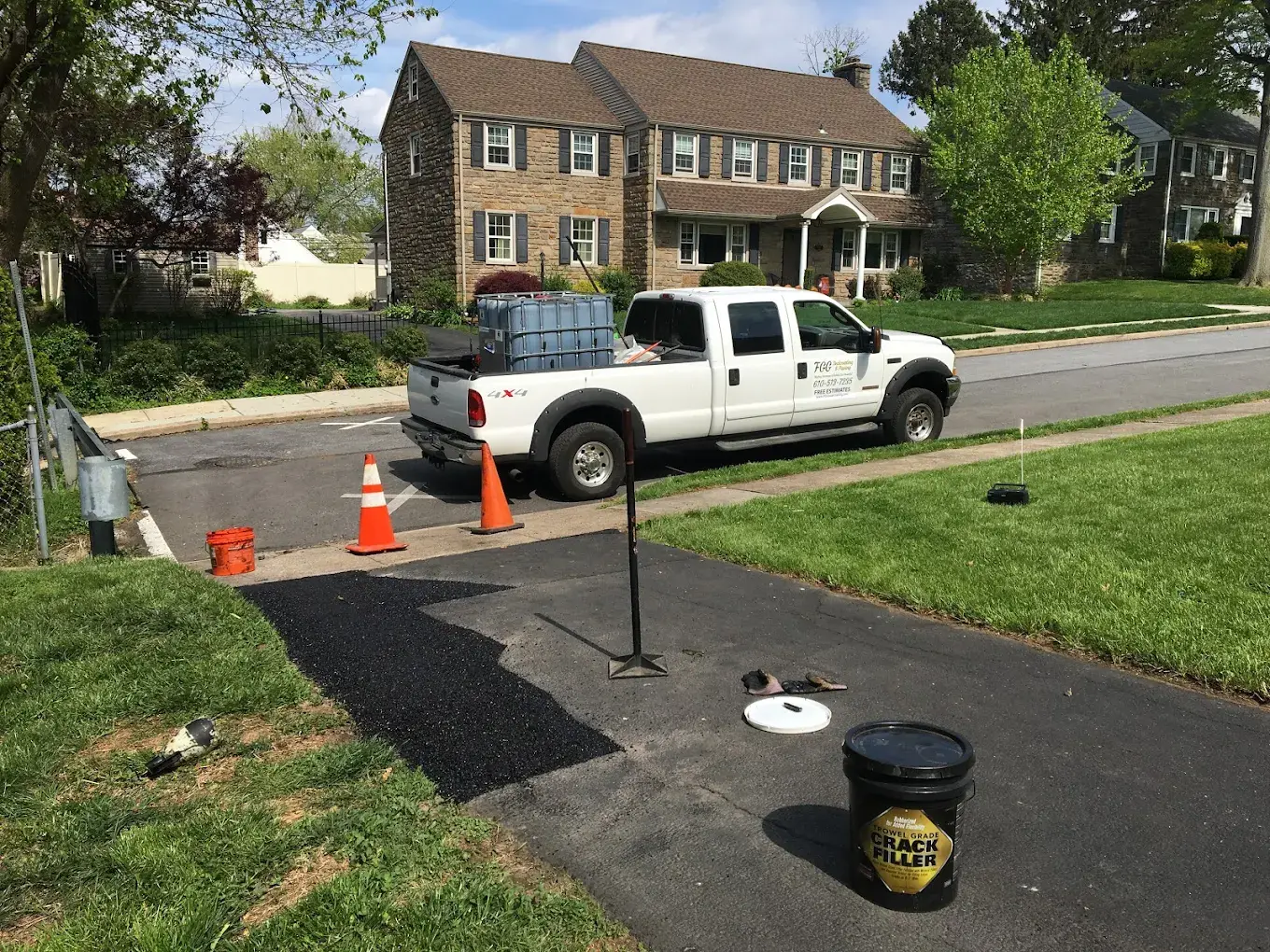 A bucket of trowel-grade crack filler and a fresh black patch at the edge of a driveway, with the FGG service truck parked on the street.