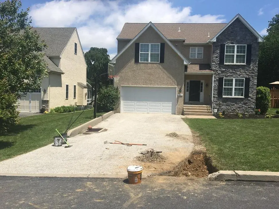 A driveway stripped to its gravel base in front of a stone-and-stucco home, with the apron cut out and tools laid on the stone.
