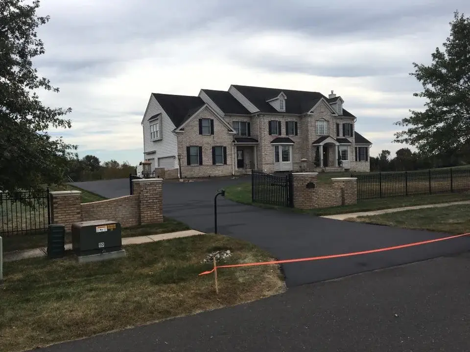 A long asphalt driveway at a large brick estate home, freshly sealed and taped off at the street with orange tape.